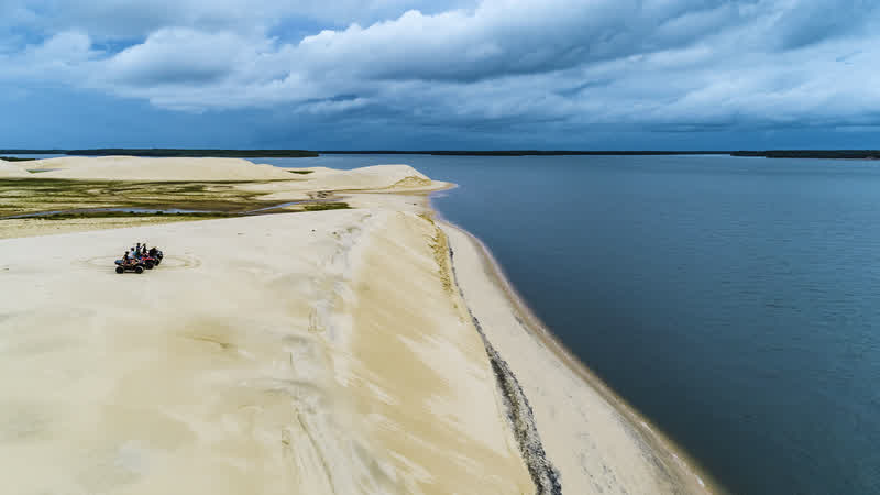Duna de areia clara terminando em água azul, com um veículo de passeio no alto