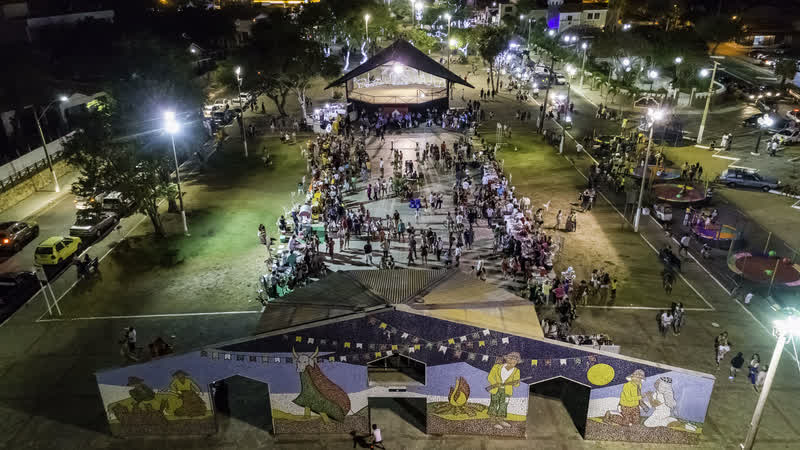 Vista aérea noturna do Quadrilhódromo, com a praça movimentada e o galpão decorado por murais coloridos